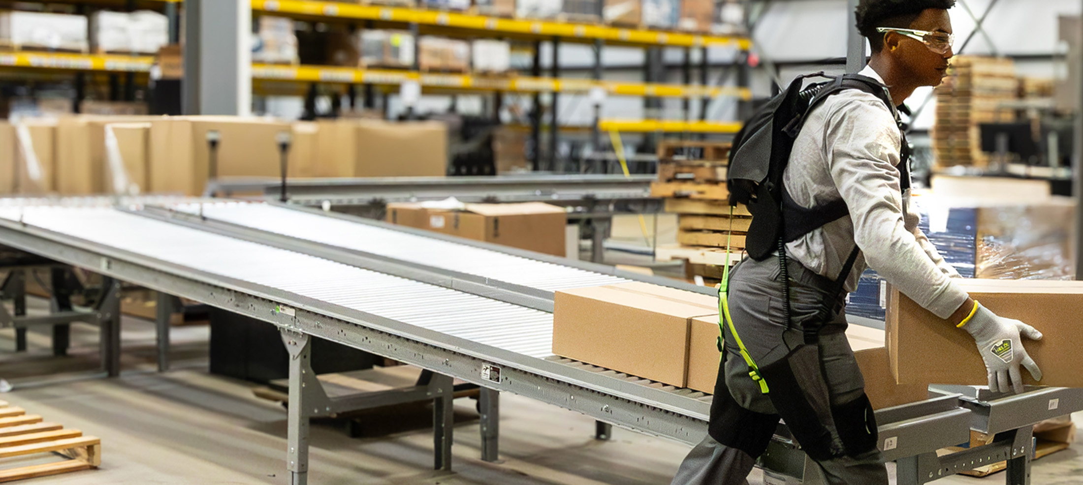 Worker carrying a box past a conveyor while wearing the SafeLift™ exosuit.